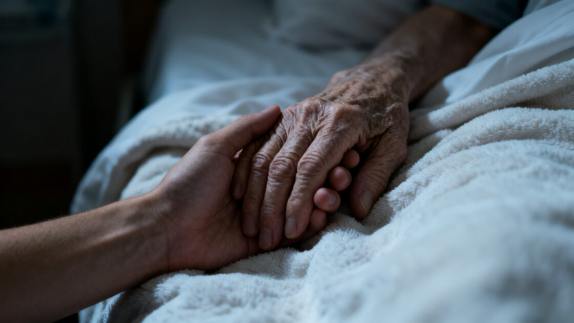Dos manos tomadas sobre la cobija de una cama, una joven y una de persona mayor