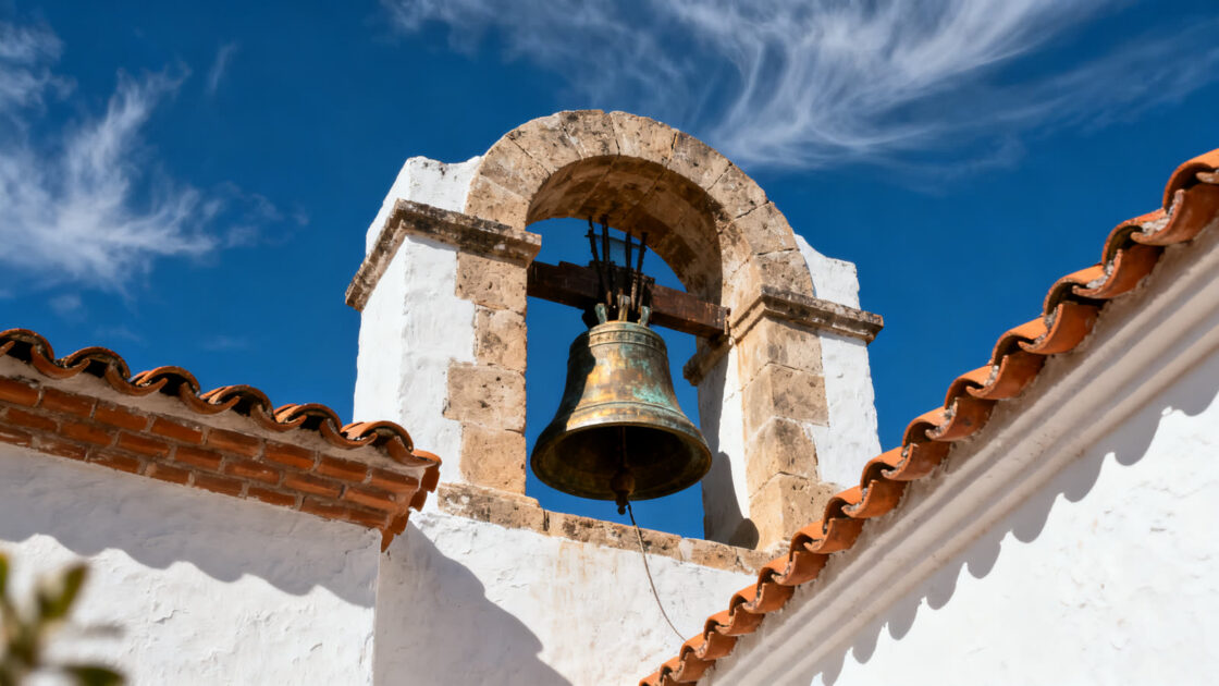 La campana de bronce de una espadaña blanca de iglesia contra un cielo azul de mediodía